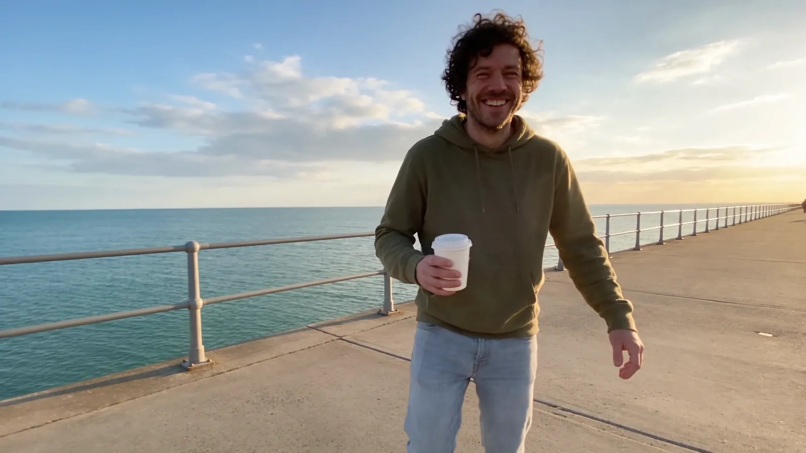 The original take: a man in an olive hoodie walks along a seaside promenade at golden hour, holding a coffee cup.
